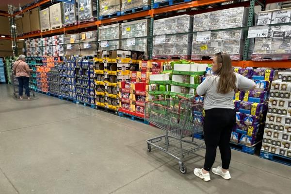 Shoppers browsing and co<em></em>nsidering purchases in a Costco warehouse, with one woman standing next to a cart full of boxes