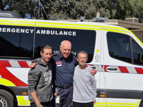 An Ambulance Community Officer smiles for the camera with his arm around two other men. They stand in front of an ambulance.