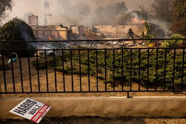 An burning and smoky residential property and structure are shown behind an iron fence, with a political sign saying 'Harris-Walz' lying on the ground in the foreground.