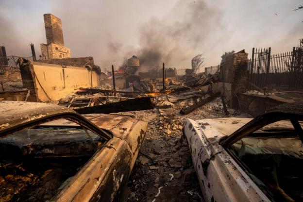 Charred vehicles are shown in the foreground on a debris-filled ground, as a burnt structure and smoke in the sky are shown.